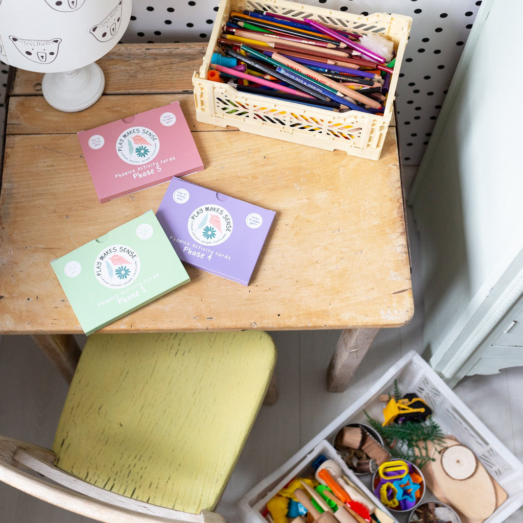 3 packs of Play Makes Sense Phonics Cards on a child's desk, next to a box of writing resources.